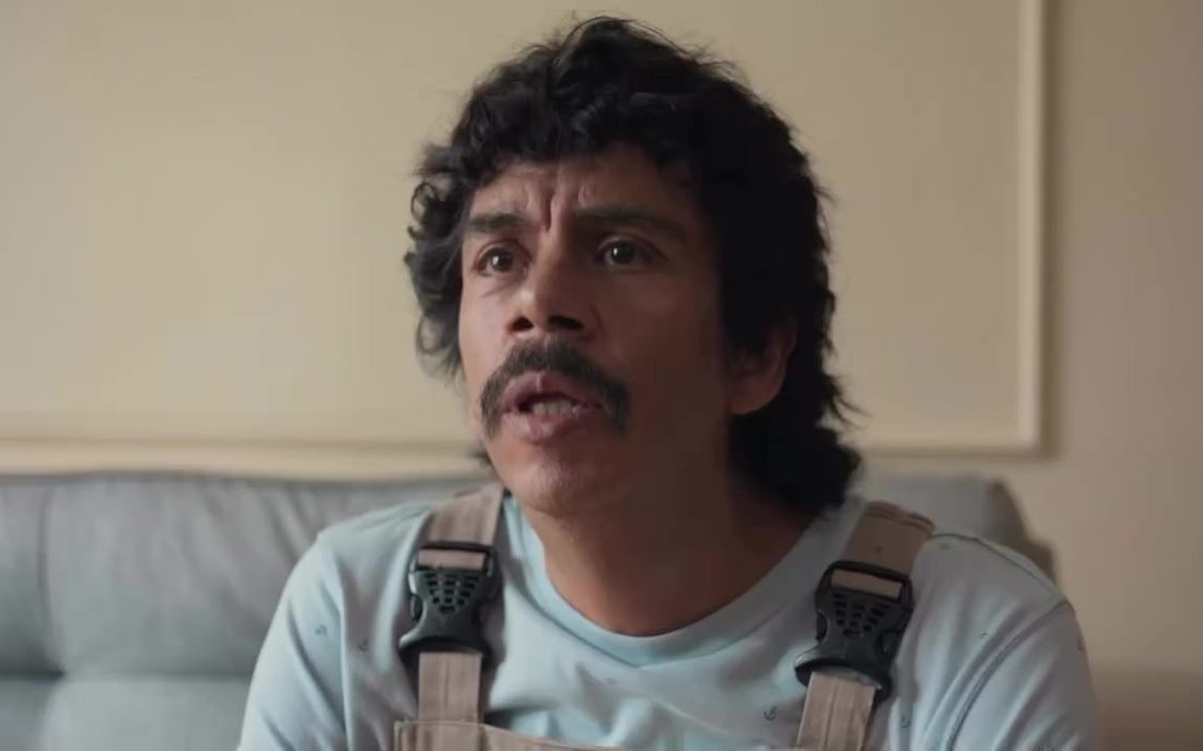 Fotograma de Bailando solo: un joven de bigote y overol habla sentado en una sala